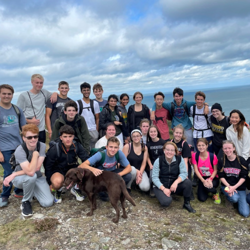 A large group of students stand at the edge of a cliff with a blue sky overhead.